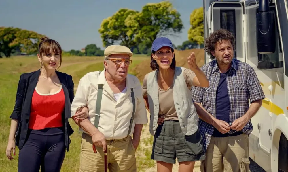 Irmãos de "Faz de conta que é Paris" (2024) de braços dados com o pai fingindo estarem em uma viagem a Paris em um trailer. Estamos diante de uma paisagem campestre.