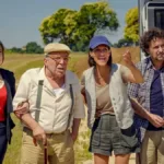 Irmãos de "Faz de conta que é Paris" (2024) de braços dados com o pai fingindo estarem em uma viagem a Paris em um trailer. Estamos diante de uma paisagem campestre.