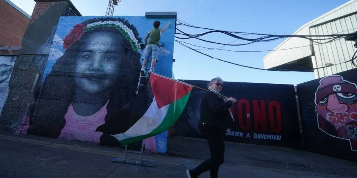 Fotografia de mural grafitado com criança sorrindo. Na rua uma mulher caminha com a bandeira da Palestina.