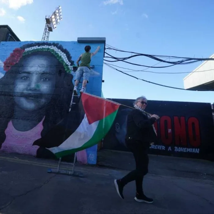 Fotografia de mural grafitado com criança sorrindo. Na rua uma mulher caminha com a bandeira da Palestina.