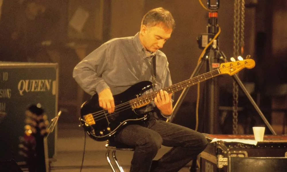 John Deacon sentado, imagem em escala sépia, tocando baixo.