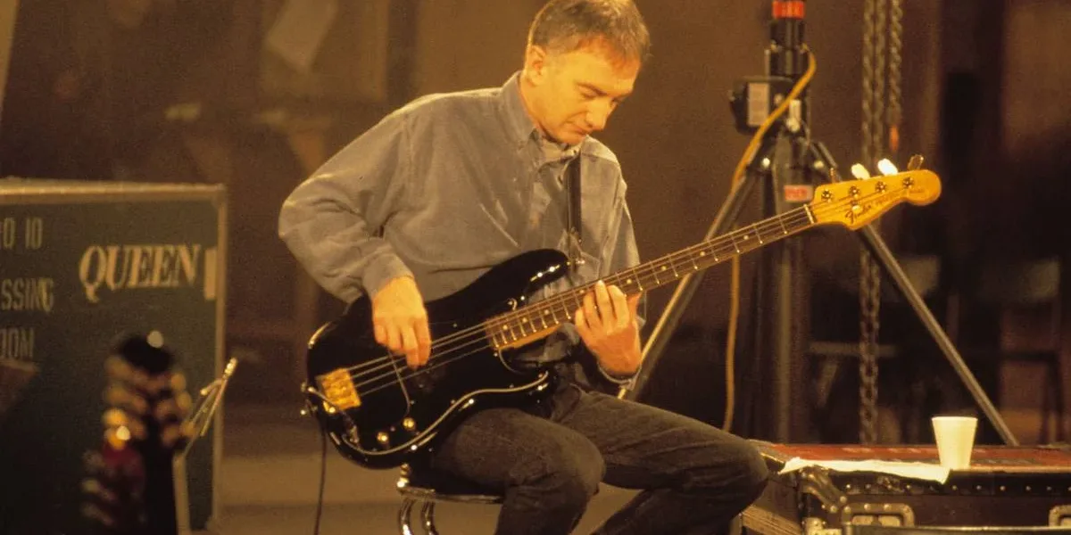 Que Fim Levou o Baixista do Queen John Deacon? John Deacon sentado, imagem em escala sépia, tocando baixo.