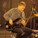 John Deacon sentado, imagem em escala sépia, tocando baixo.