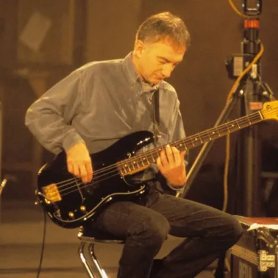 John Deacon sentado, imagem em escala sépia, tocando baixo.