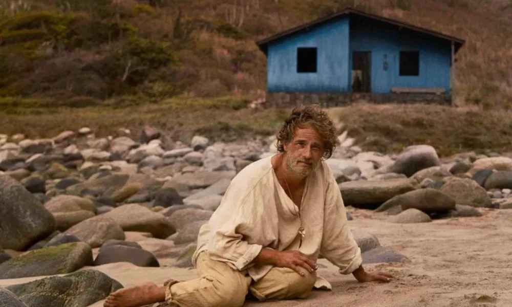 Rodrigo Santoro como Crisóstomo, protagonista do longa "O Filho de Mil Homens", destaque da Mostra Internacional de Cinema de São Paulo de 2025. Ele está maltrapilho, roupas bege, sentado/caído no chão arenoso e com pedras ao fundo. Há uma paisagem com grama em tons marrom ao fundo e uma casinha azul.