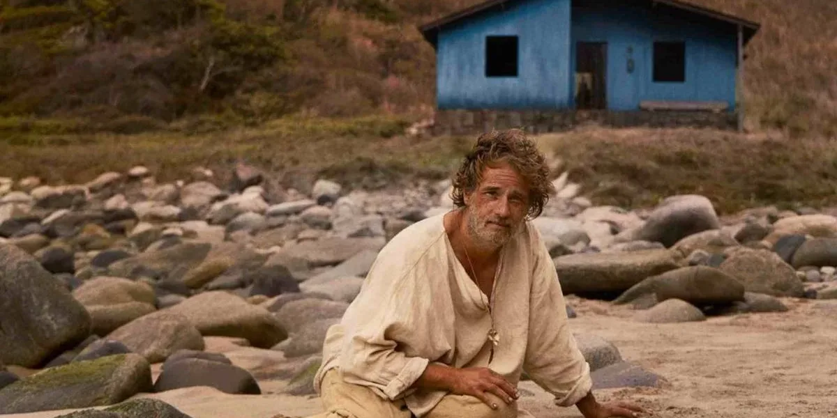 Rodrigo Santoro como Crisóstomo, protagonista do longa "O Filho de Mil Homens", destaque da Mostra Internacional de Cinema de São Paulo de 2025. Ele está maltrapilho, roupas bege, sentado/caído no chão arenoso e com pedras ao fundo. Há uma paisagem com grama em tons marrom ao fundo e uma casinha azul.