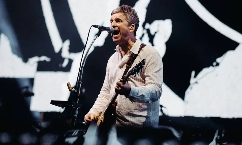 Noel Gallagher, camiseta social branca levemente rosada, tocando guitarra em show dia 16 de agosto de 2025 em Dublin. Imagem colorida.