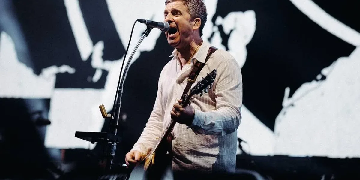 Noel Gallagher, camiseta social branca levemente rosada, tocando guitarra em show dia 16 de agosto de 2025 em Dublin. Imagem colorida.