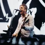 Noel Gallagher, camiseta social branca levemente rosada, tocando guitarra em show dia 16 de agosto de 2025 em Dublin. Imagem colorida.