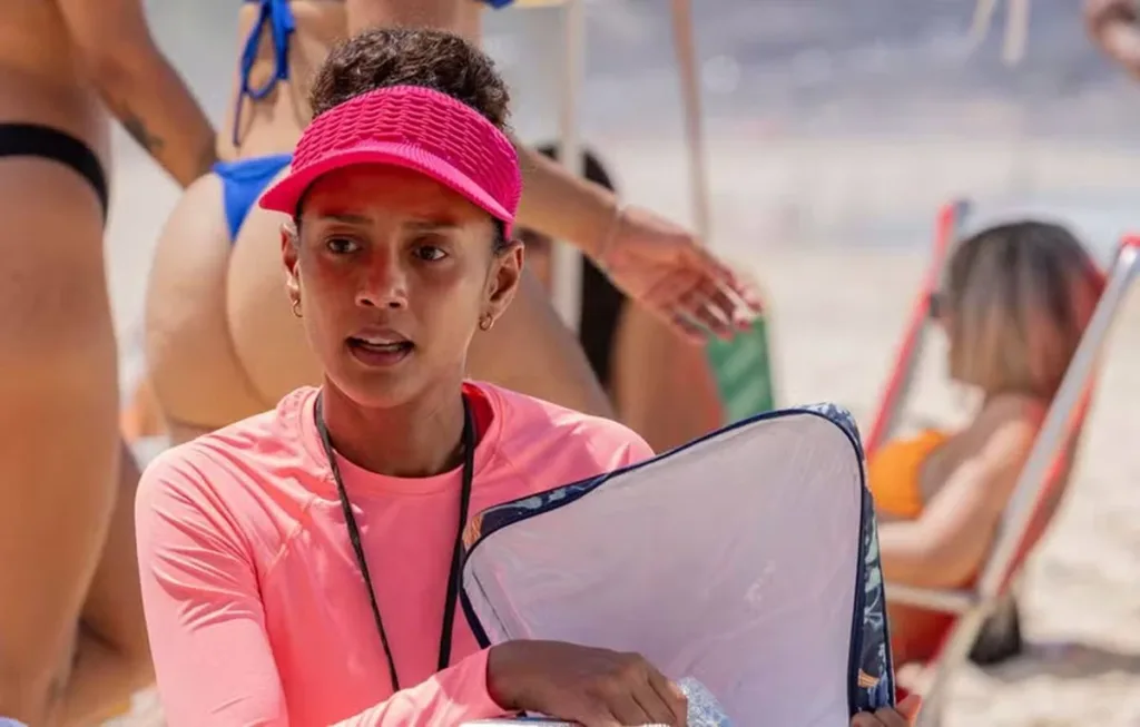 Vale Tudo | Entenda a Sequência de Furos Que Está Indignando os Telespectadores 1 Taís Araújo como Raquel Acioli, de viseira e roupa rosa vendendo sanduíches na praia após virada na novela "Vale Tudo" de 2025.