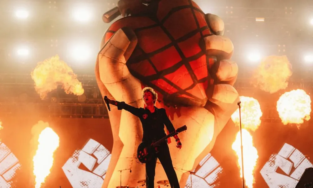 Billie Joe Armstrong com mão direita levantada com microfone à plateia, em foto centralizado no palco Skyline do The Town 2025. Ao fundo há pirotecnia e uma representação da capa do álbum American Idiot esculpida.