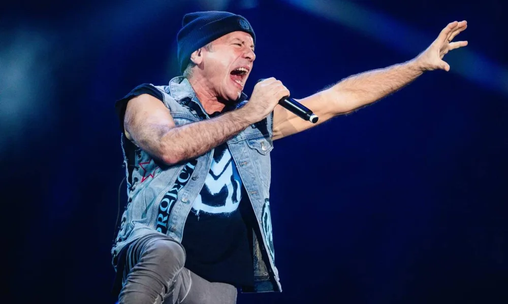 Bruce Dickinson, jaqueta de couro, touca preta, segurando microfone na mão direita e com a mão direita estendida e aberta em garra, cantando com boca bem aberta em foto em seu show no Palco Skyline no The Town 2025.
