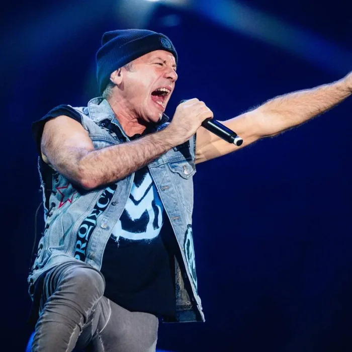 Bruce Dickinson, jaqueta de couro, touca preta, segurando microfone na mão direita e com a mão direita estendida e aberta em garra, cantando com boca bem aberta em foto em seu show no Palco Skyline no The Town 2025.