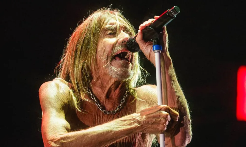 Cãmera focada em Iggy Pop, sutilmente inclinado ao microfone, que segura com as duas mãos, em luz baixa e fundo preto no palco The One no The Town 2025.