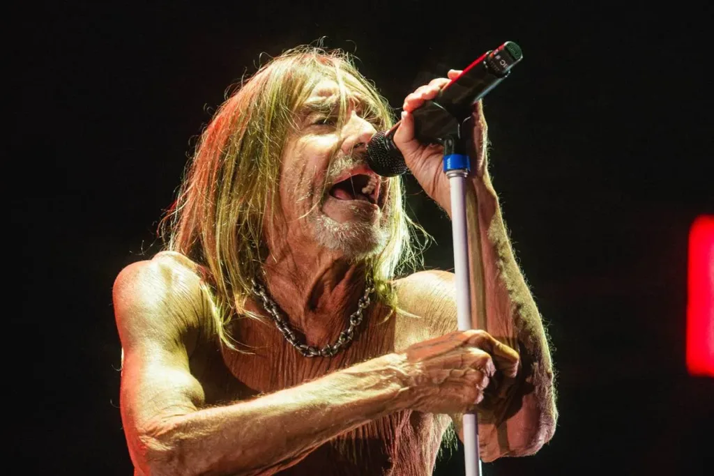 The Town 2025 | Os 10 Melhores Shows do Primeiro Final de Semana 6 Cãmera focada em Iggy Pop, sutilmente inclinado ao microfone, que segura com as duas mãos, em luz baixa e fundo preto no palco The One no The Town 2025.