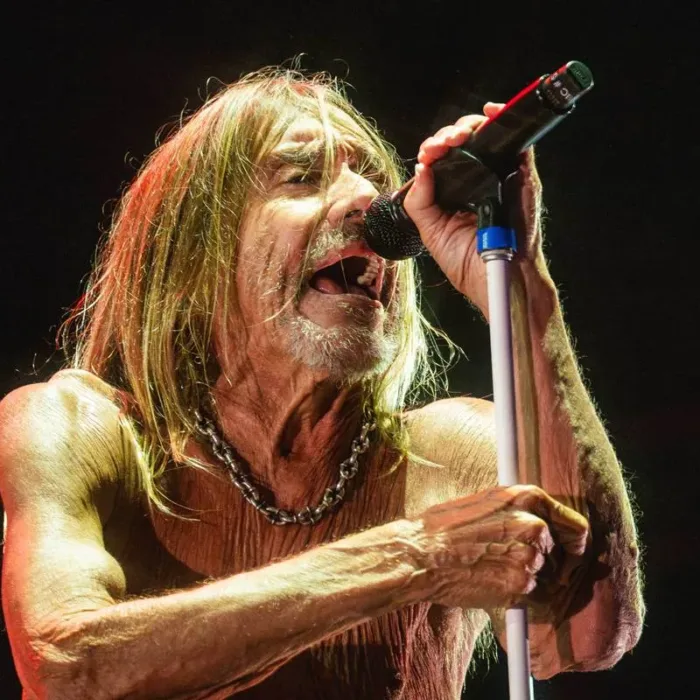 Cãmera focada em Iggy Pop, sutilmente inclinado ao microfone, que segura com as duas mãos, em luz baixa e fundo preto no palco The One no The Town 2025.