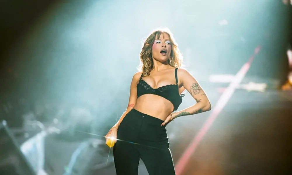 Luisa Sonza no palco The One do The Town 2025. Ela está com mão na cintura, vestindo jeans e sutiã pretos, meio inclinada para sua esquerda, segurando o microfone na mão direita e de boca aberta. Está com maquiagem forte, pegada no blush e com azul forte ao redor dos olhos.