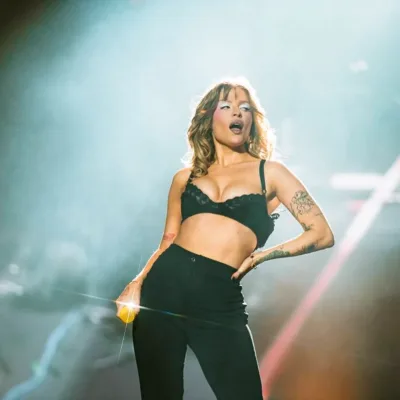 Luisa Sonza no palco The One do The Town 2025. Ela está com mão na cintura, vestindo jeans e sutiã pretos, meio inclinada para sua esquerda, segurando o microfone na mão direita e de boca aberta. Está com maquiagem forte, pegada no blush e com azul forte ao redor dos olhos.