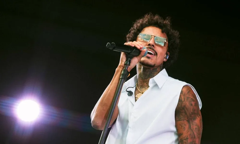 Livinho com um colete branco, segurando o microfone com a mão direita, e rosto levemente inclinado no sentido oposto, de óculos no palco Factory do sábado do The Town 2025.