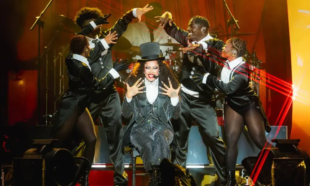 Gloria Groove sentada em um banquinho no seu show no palco Skyline, que imita um cabaré. Tanto ela quanto os quatro dançarinos a sua volta estão com trajes de cabaré em preto e branco. Gloria mostrando as mãos ao público com um sorriso no rosto enquanto os dançarinos balançam as mãos em torno dela, como se fizessem suspense.