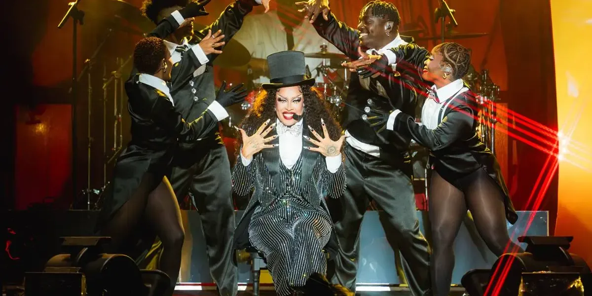 Gloria Groove sentada em um banquinho no seu show no palco Skyline, que imita um cabaré. Tanto ela quanto os quatro dançarinos a sua volta estão com trajes de cabaré em preto e branco. Gloria mostrando as mãos ao público com um sorriso no rosto enquanto os dançarinos balançam as mãos em torno dela, como se fizessem suspense.