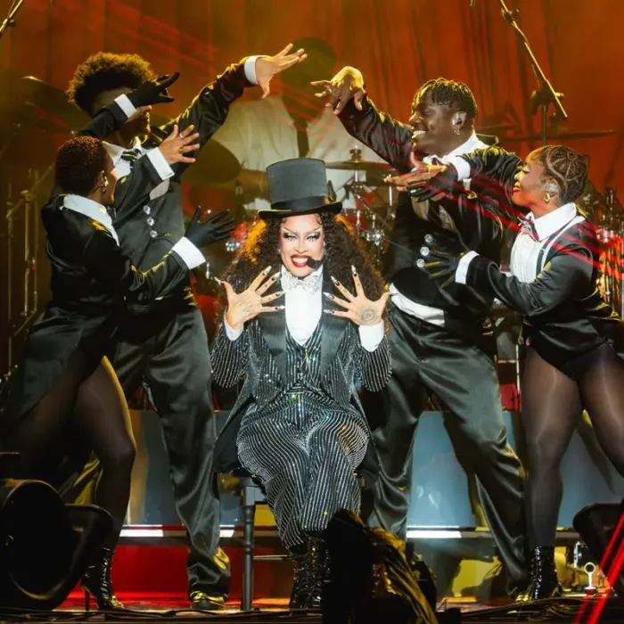 Gloria Groove sentada em um banquinho no seu show no palco Skyline, que imita um cabaré. Tanto ela quanto os quatro dançarinos a sua volta estão com trajes de cabaré em preto e branco. Gloria mostrando as mãos ao público com um sorriso no rosto enquanto os dançarinos balançam as mãos em torno dela, como se fizessem suspense.