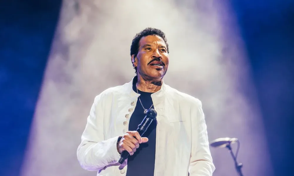 Lionel Richie segurando o microfone na mão direita na altura da barriga, próximo ao corpo, de terno branco, com um sorrisinho durante sua apresentção no segundo sábado de The Town 2025.