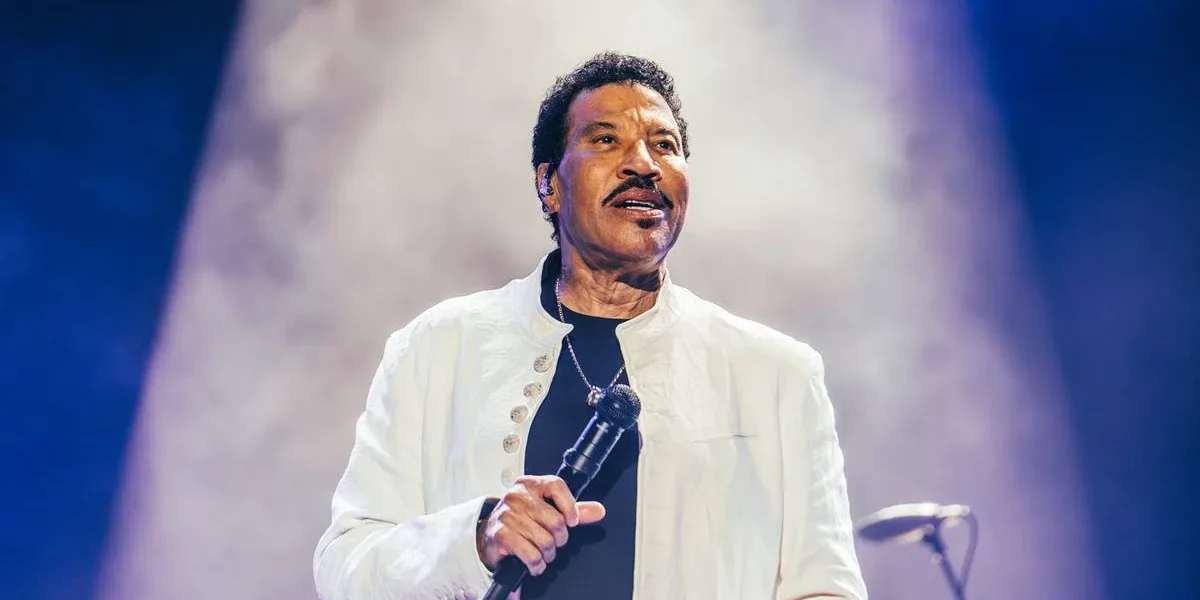 Lionel Richie segurando o microfone na mão direita na altura da barriga, próximo ao corpo, de terno branco, com um sorrisinho durante sua apresentção no segundo sábado de The Town 2025.