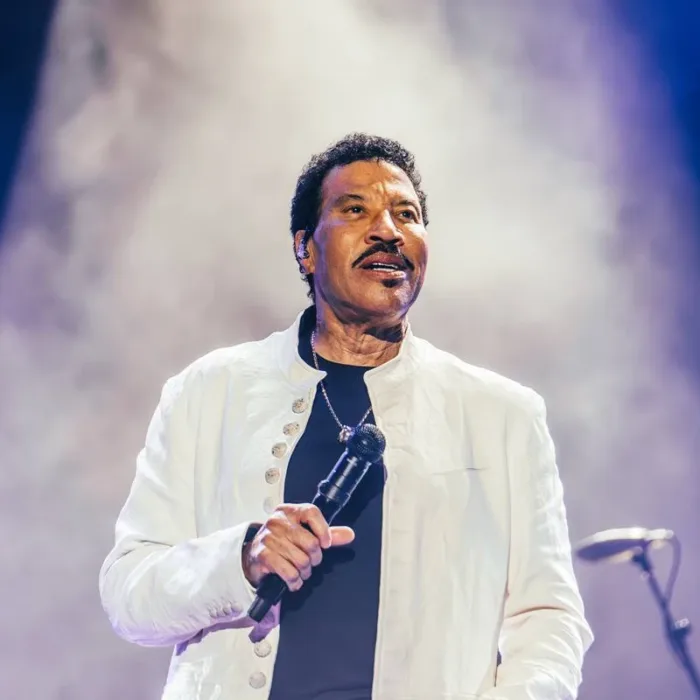 Lionel Richie segurando o microfone na mão direita na altura da barriga, próximo ao corpo, de terno branco, com um sorrisinho durante sua apresentção no segundo sábado de The Town 2025.