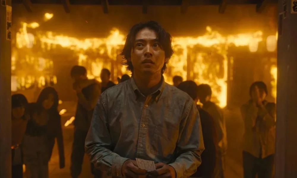 Arisu no segundo episódio da terceira temporada de "Alice in Borderland". Roupa social, no meio de uma multidão em um ambiente em chamas.