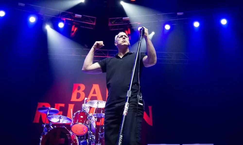 Greg Gaffin do Bad Religion em palco em show com co-headline do Social Distortion. Ao fundo há uma bateria e o logo da banda em telão. Paleta de cores preta luzes azuis-arroxeadas.
