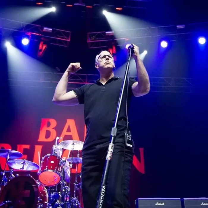 Greg Gaffin do Bad Religion em palco em show com co-headline do Social Distortion. Ao fundo há uma bateria e o logo da banda em telão. Paleta de cores preta luzes azuis-arroxeadas.