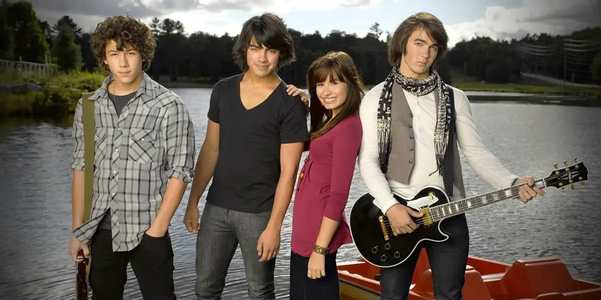 Camp Rock 3 | Tudo o Que Já Sabemos do Novo Filme Nick e Joe Jonas, Demi Lovato, e Kevin Jonas, nessa ordem da esquerda para a direita, posando para foto promocional do primeiro filme "Camp Rock", caracterizados para seus personagens do filme e na frente de um lago.