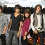 Nick e Joe Jonas, Demi Lovato, e Kevin Jonas, nessa ordem da esquerda para a direita, posando para foto promocional do primeiro filme "Camp Rock", caracterizados para seus personagens do filme e na frente de um lago.
