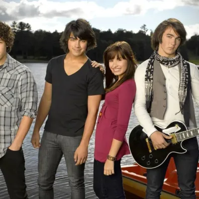 Nick e Joe Jonas, Demi Lovato, e Kevin Jonas, nessa ordem da esquerda para a direita, posando para foto promocional do primeiro filme "Camp Rock", caracterizados para seus personagens do filme e na frente de um lago.
