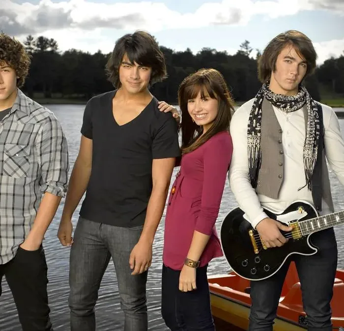 Nick e Joe Jonas, Demi Lovato, e Kevin Jonas, nessa ordem da esquerda para a direita, posando para foto promocional do primeiro filme "Camp Rock", caracterizados para seus personagens do filme e na frente de um lago.