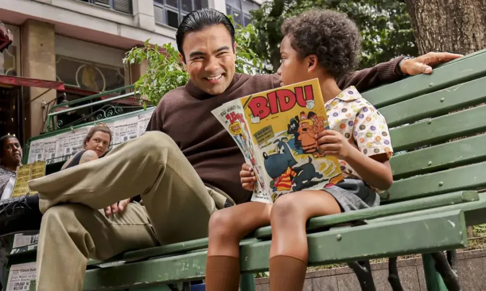 Mauro Sousa, filho de Maurício de Sousa, interpretando o pai mais jovem, sentado em um banco de madeira pintado em verde, ao lado de um garoto lendo um gibi do Bidu.
