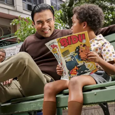 Mauro Sousa, filho de Maurício de Sousa, interpretando o pai mais jovem, sentado em um banco de madeira pintado em verde, ao lado de um garoto lendo um gibi do Bidu.