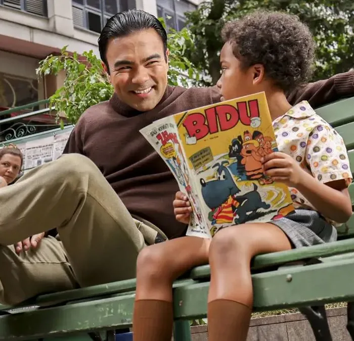 Mauro Sousa, filho de Maurício de Sousa, interpretando o pai mais jovem, sentado em um banco de madeira pintado em verde, ao lado de um garoto lendo um gibi do Bidu.