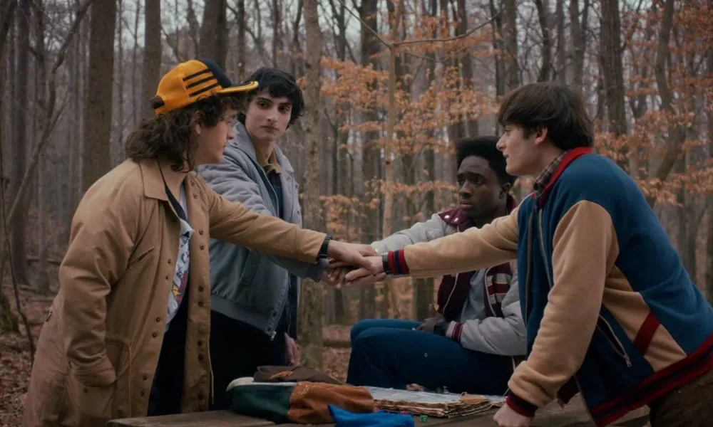 Os quatro protagonistas masculinos de Stranger Things dando as mãos em uma rodinha no primeiro episódio da última temporada, em um bosque que parece estar no outono.