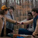 Os quatro protagonistas masculinos de Stranger Things dando as mãos em uma rodinha no primeiro episódio da última temporada, em um bosque que parece estar no outono.