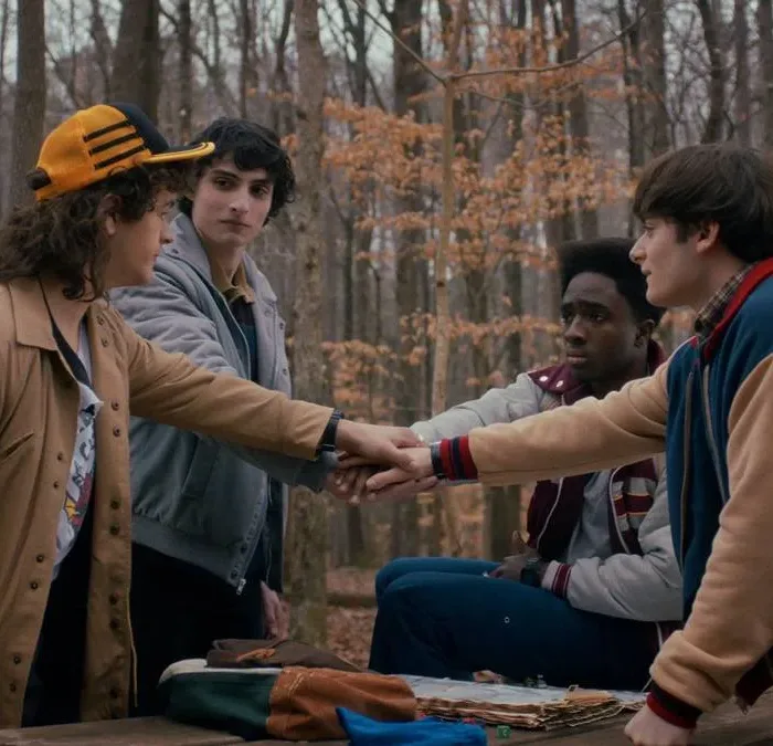 Os quatro protagonistas masculinos de Stranger Things dando as mãos em uma rodinha no primeiro episódio da última temporada, em um bosque que parece estar no outono.