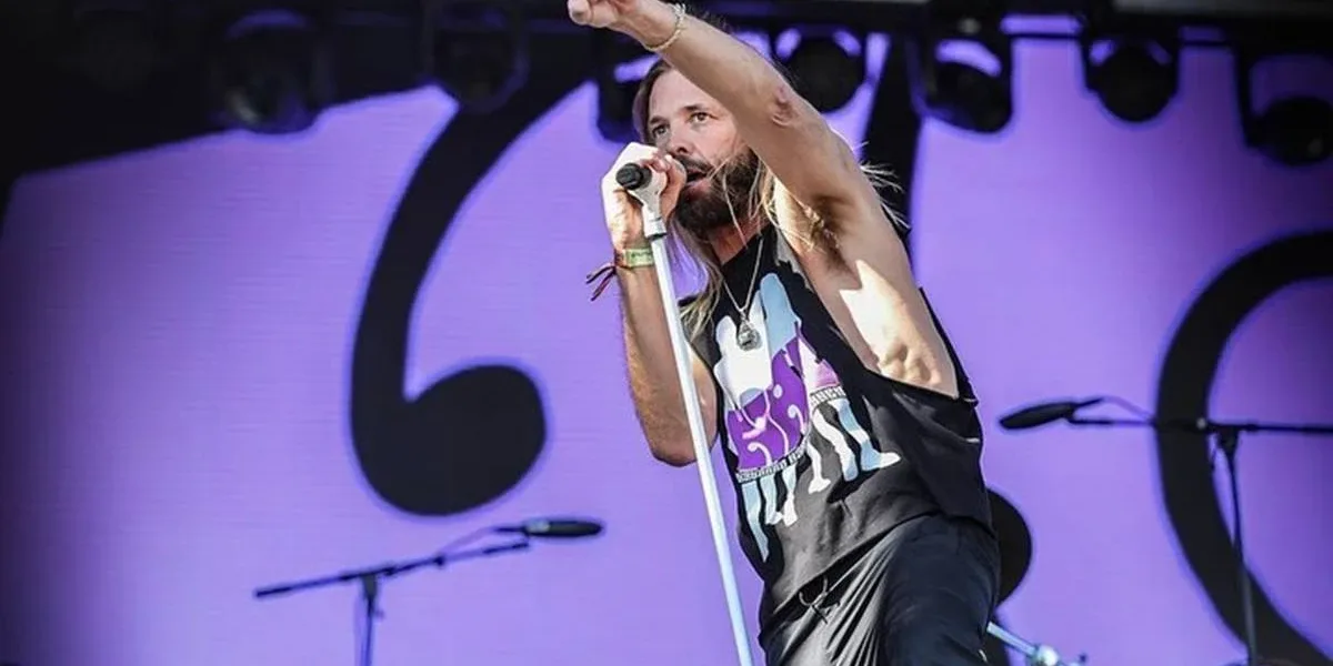 Taylor Hawkins em ângulo plongée, de regata preta e jeans, apontando com o indicador para a plateia com o braço direito levantado e o esquero segurando o microfone em apresentação.