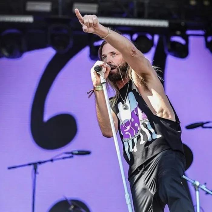 Taylor Hawkins em ângulo plongée, de regata preta e jeans, apontando com o indicador para a plateia com o braço direito levantado e o esquero segurando o microfone em apresentação.