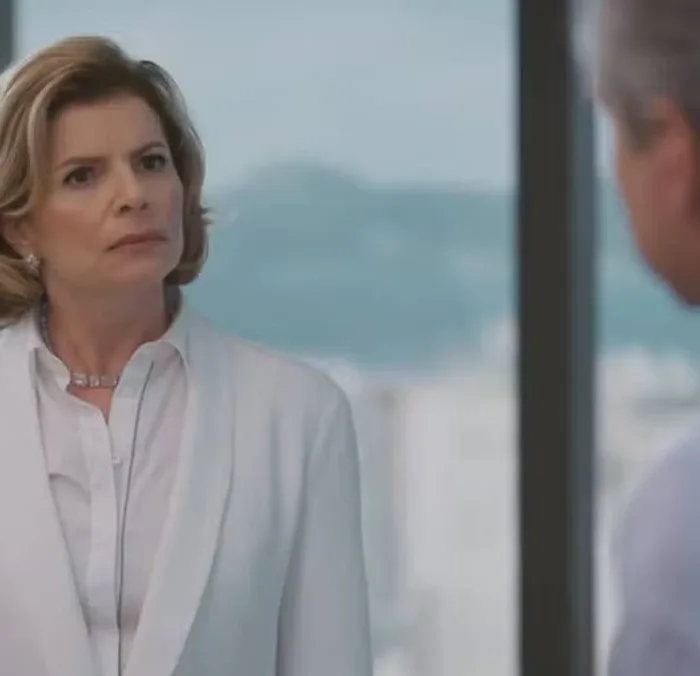 Débora Bloch como Odete Roitman em Vale Tudo, de roupa social branca, encarando irritada Marco Aurélio (Alexandre Nero) que está de costas para a tela, de frente para nós.