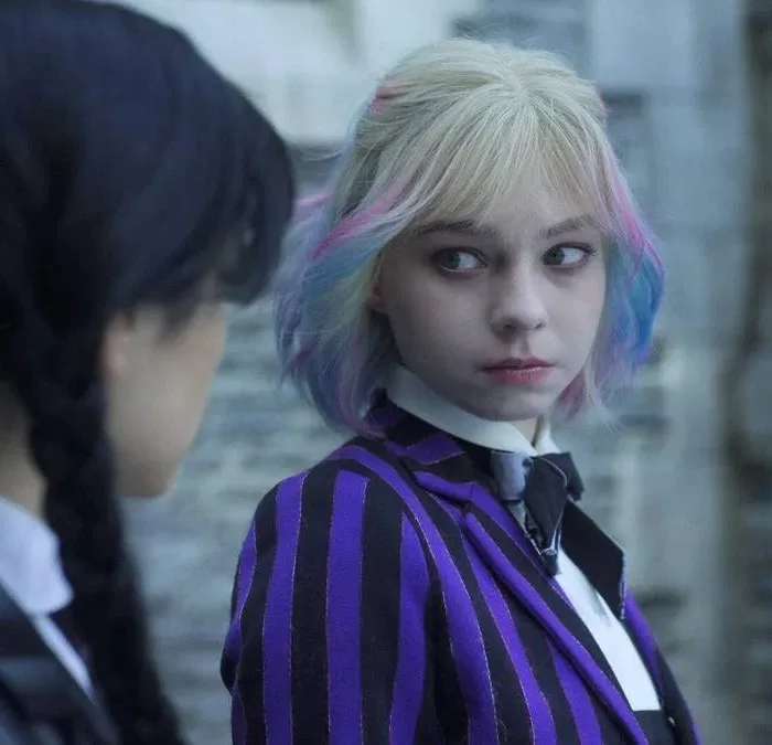 Jenna Ortega de lado como Wandinha, e Emma Myers sutilmente virada para o lado com o uniforme de Nunca Mais, encarando-a, como a personagem Enid Sinclair. Wandinha e Enid estão com corpos trocados, razão pela qual Enid é na verdade Wandinha e está a encarando profundamente.