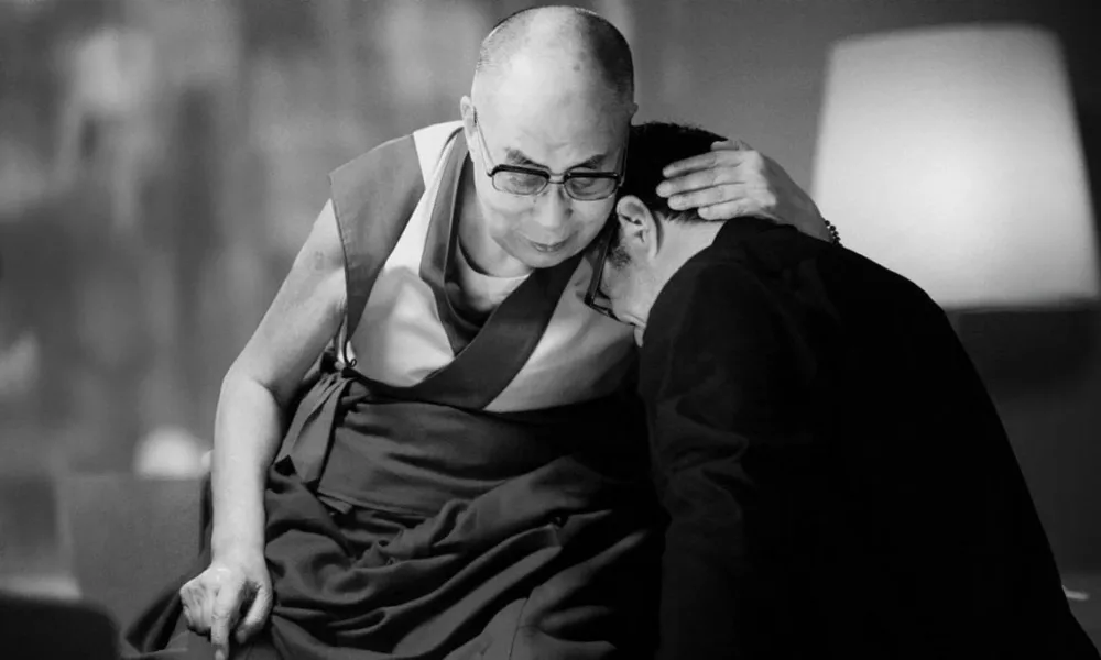 Foto em preto e branco com Dalai Lama abraçando um homem em seus ombros. Foto do filme "A Sabedoria da Felicidade".