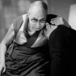 Foto em preto e branco com Dalai Lama abraçando um homem em seus ombros. Foto do filme "A Sabedoria da Felicidade".