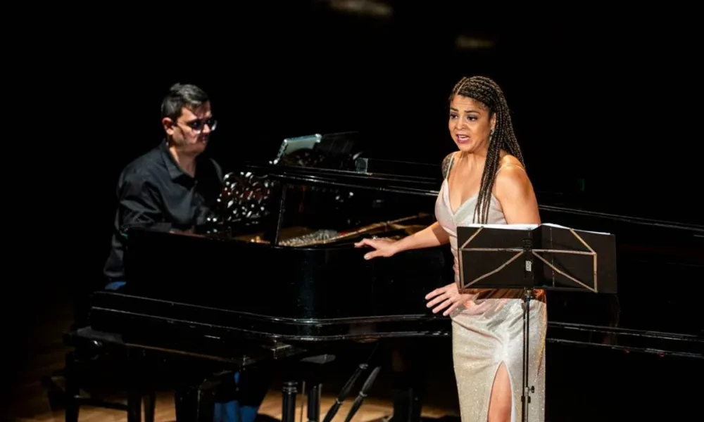 Chiara Santoro cantando em recital "As Brasileiras", partitura em sua frente e ela voltada ao público. Atrás dela está o pianista Silas Barbosa.