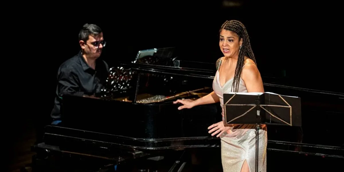 Chiara Santoro cantando em recital "As Brasileiras", partitura em sua frente e ela voltada ao público. Atrás dela está o pianista Silas Barbosa.